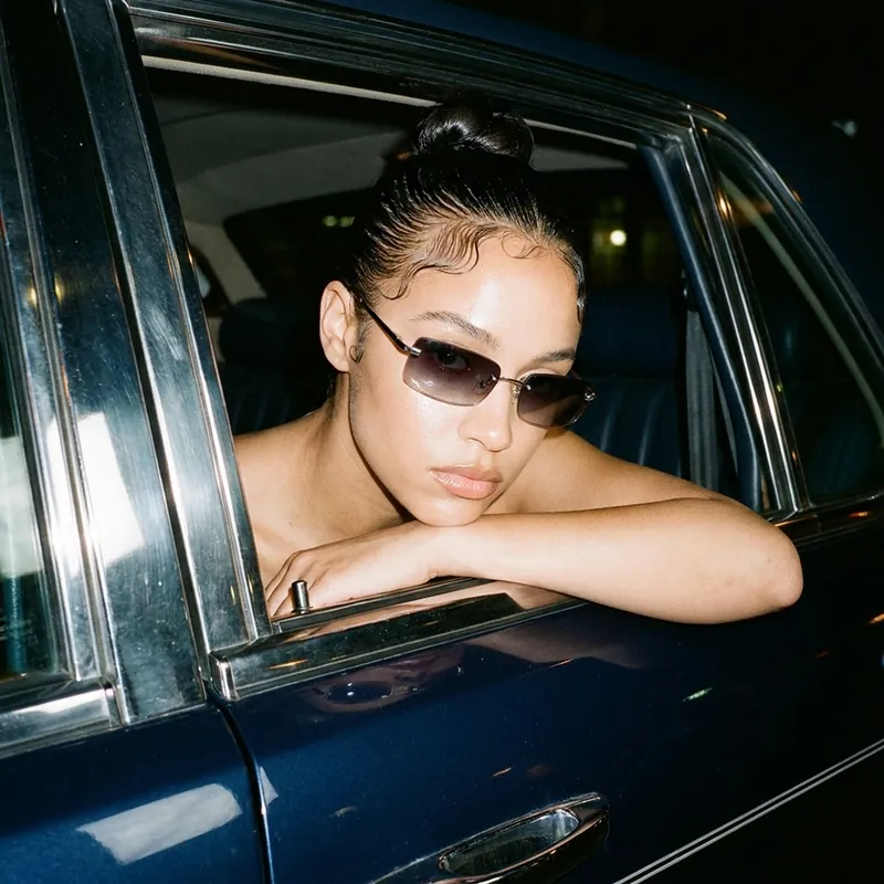 Une femme portant des lunettes de soleil étroites appuyée contre la portière d'une voiture vintage sombre la nuit