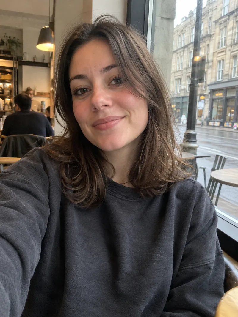 The AI-generated stand-in selfie used for every test: a woman with shoulder-length brown hair in a dark grey sweatshirt, smiling in a cafe by a rainy street window