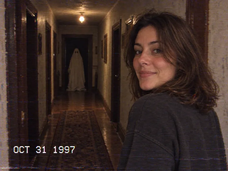 ChatGPT VHS camcorder still: the woman looking back over her shoulder in a dim hallway with one ceiling bulb, a white bedsheet ghost at the far end, and the timestamp OCT 31 1997